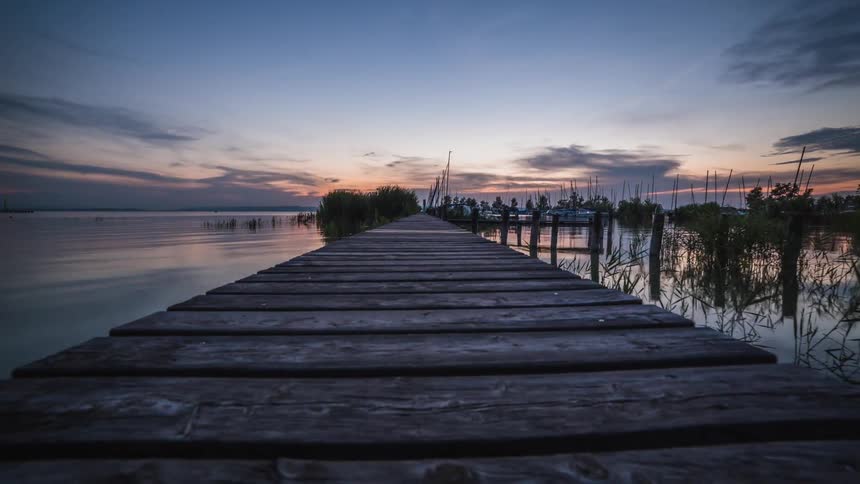 A small sailboat sails past a wooden dock and a person walks past at sunset