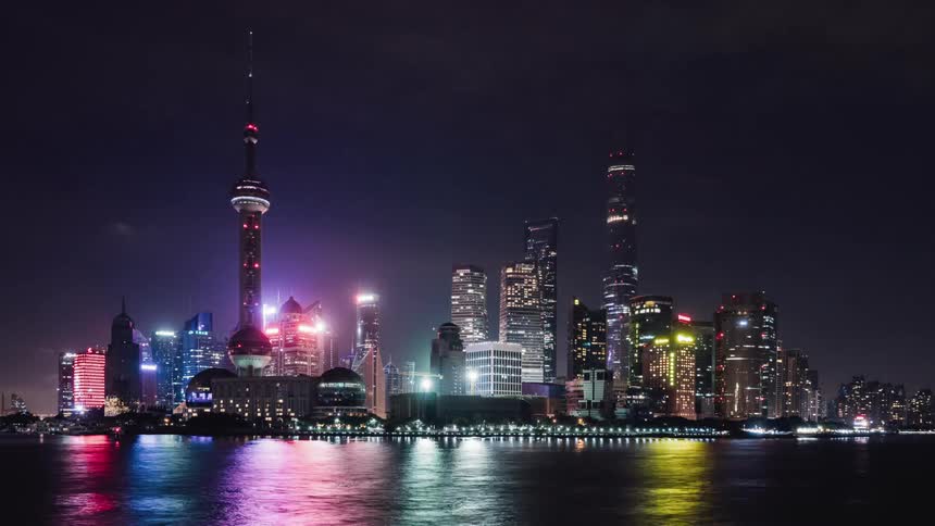 Illuminated buildings in a city sit next to the river as boats travel by