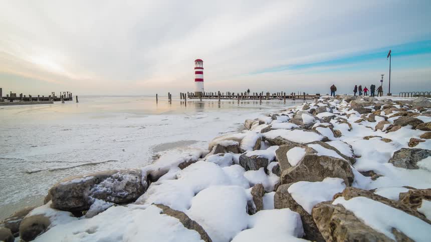 People walk along a rocky snowy jetty and pier towards a red and white lighthouse