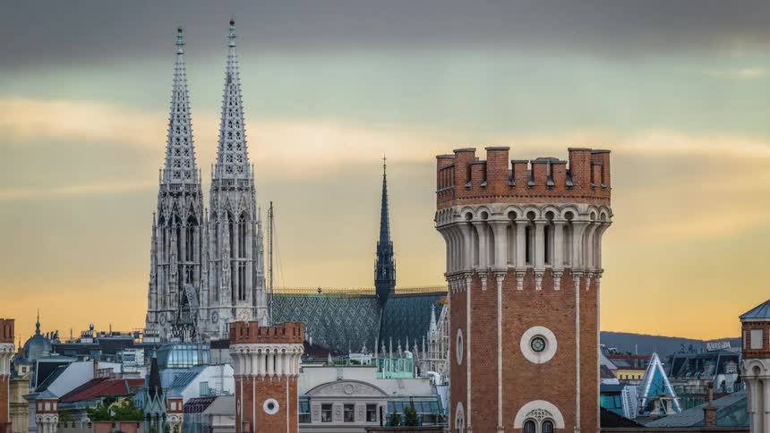 On a overcast day the spires of a church with classical architecture stands behind two brick towers