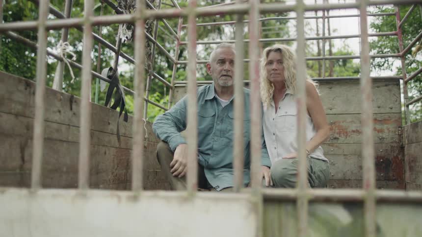 Man in blue shirt and women in white shirt and green pants sit behind bars in a wooden enclosure with trees in the background as a rifle is pointed at them