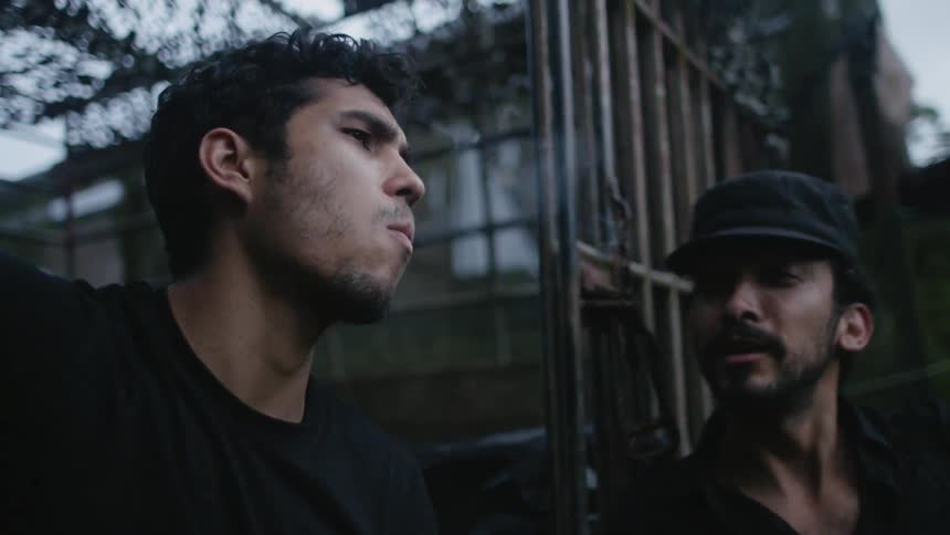 A man smokes a cigarette while standing with another man next to a building