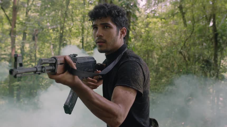 A gun wielding man slowly walks through a forest while leaves and debris fall on him