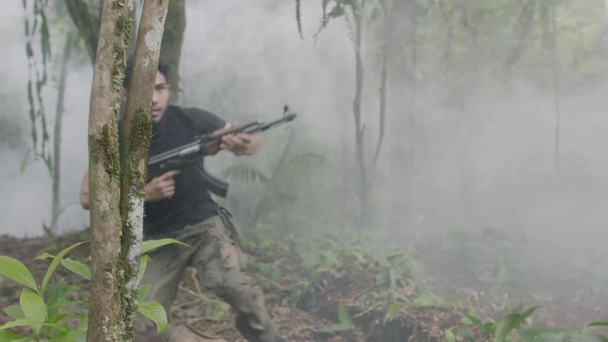 Several men can dressed in black shirts and camouflage pants are running through a smoke filled jungle while they are carrying machine guns and explosions are happening around them