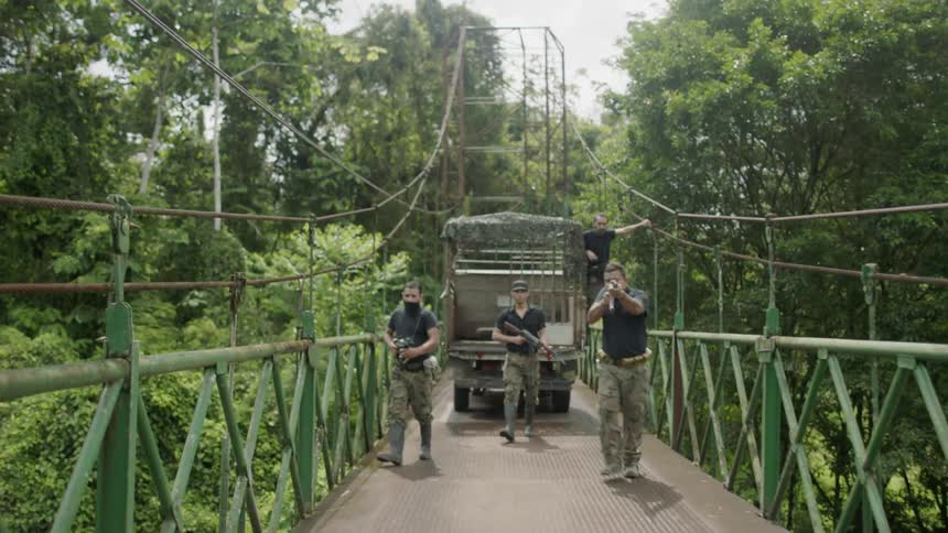 A group of mean wearing camouflage pants and carrying machine guns walk along a suspended bridge in a jungle area and walk threateningly towards a stopped vehicle that is also on the bridge