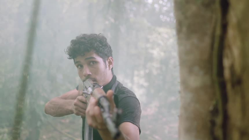 Man holding guns stands in a forest behind a tree surrounded by the mist peeping out