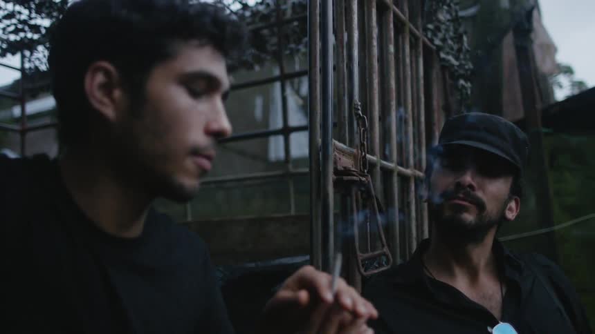 Two men talk and share a cigarette near a cage