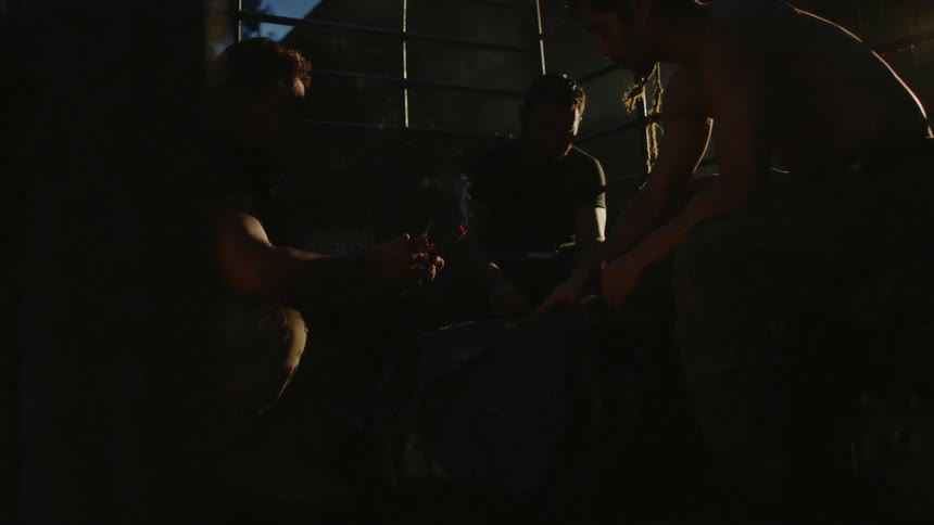 Three men in military gear smoke cigars and play cards in a camp after sunset
