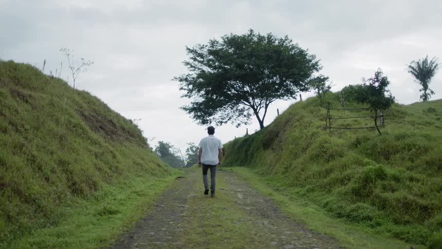 A man in a shortsleeved shirt and pants walks down a dirt road with a grassy hill on either side and a large tree up ahead