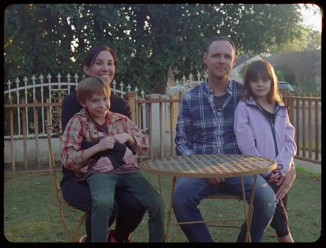 A family of four comprised of a mother father son and daughter sit at a bistro table in a yard posing for a picture