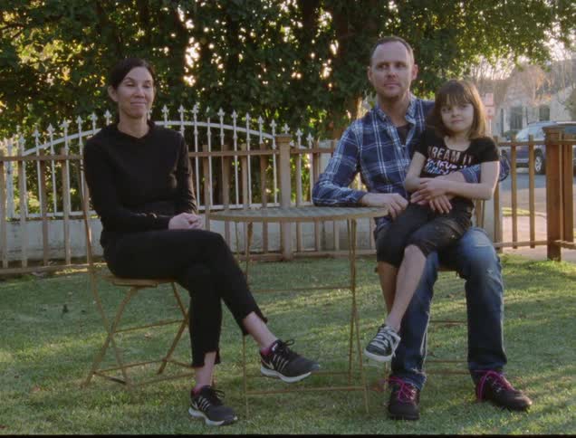 A couple sit on a lawn at a table and talk to one another while the father balances his daughter on his lap
