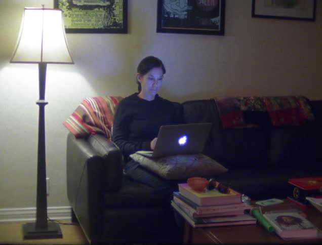 A woman sits on a couch working on her laptop propped on her lap with a pillow