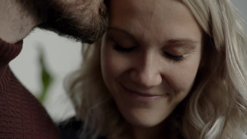A blonde haired woman smiles and looks downward as she is kissed on the side of her forehead by a tall bearded man