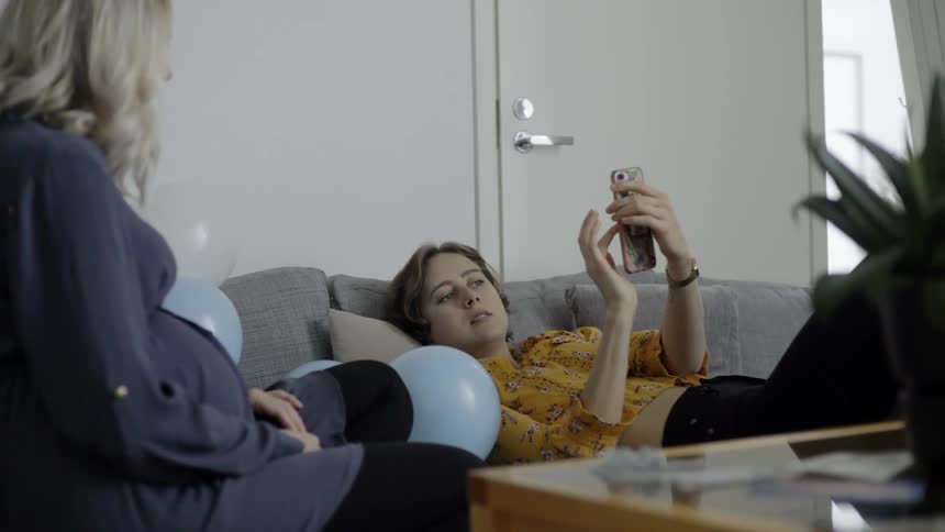 A woman laying on a couch is showing another woman something on her smart phone