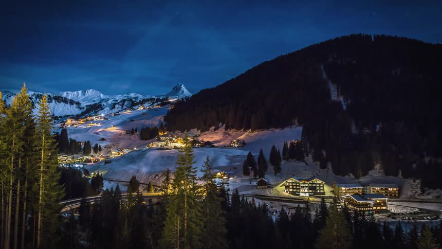 Cars drive down a winding road through a mountainside town covered in snow