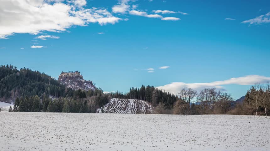 Clouds move over a snow covered field with a forest in the background