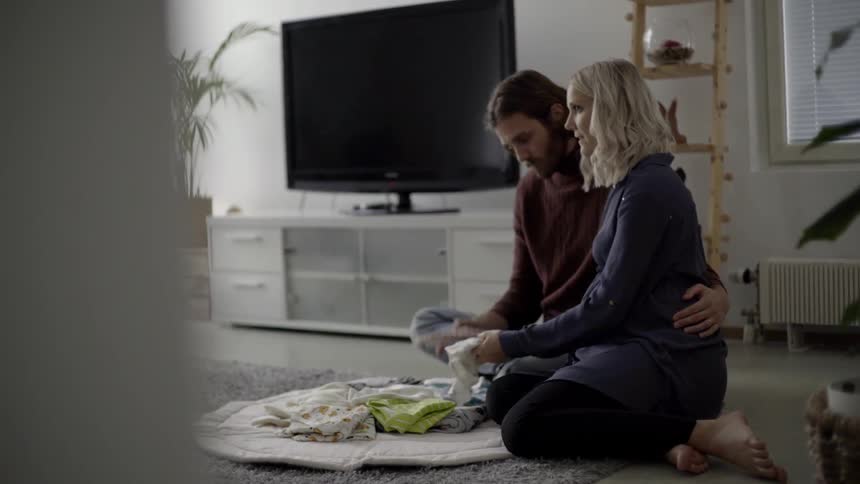 A young couple look at baby clothes while sitting on a rug in their living room