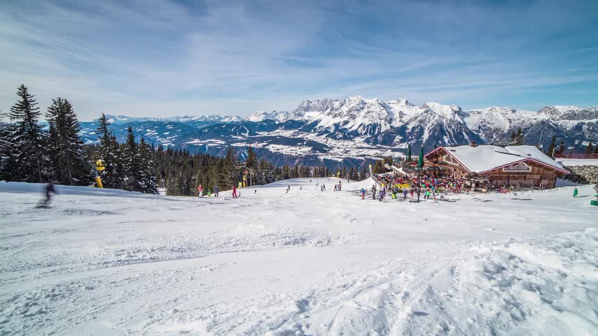 Skiers and snowboards ski down a gentle slope towards the crowds gathered at a ski lodge on a sunny day