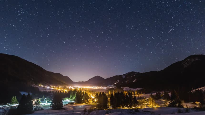 Shooting stars pass over glowing village and mountains landscape