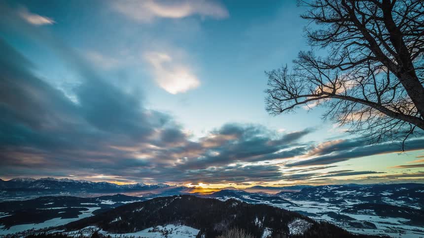 Clouds move across the blue sky over bare tree branches and snow capped elevations