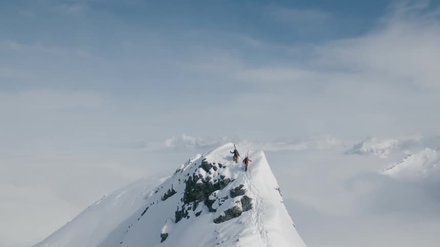 Two people climb to the top of a snow covered mountain peak