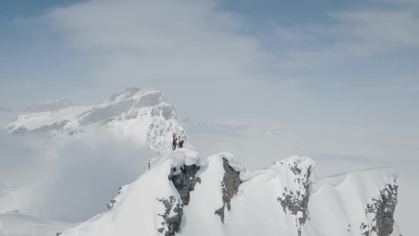 Two people stand atop a snow covered mountain top high above the group below and surrounded by other impressive mountains