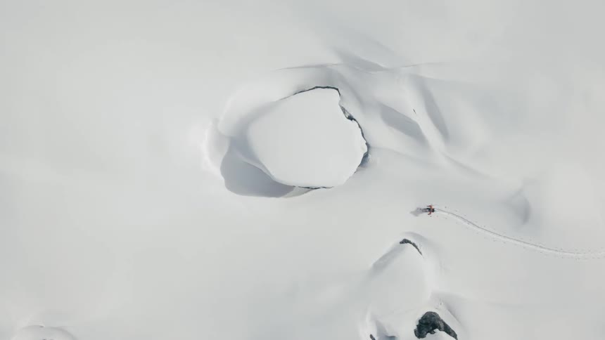 An aerial view of a person hiking in deep snow on a rocky terrain while wearing skis