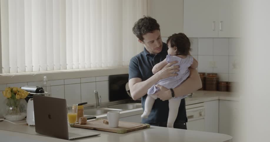 A standing in a kitchen holding a baby while using an apple laptop