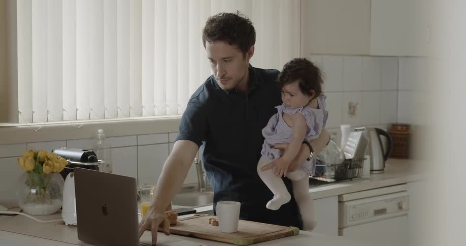 Man holding baby drinks coffee and uses laptop in the kitchen during daylight