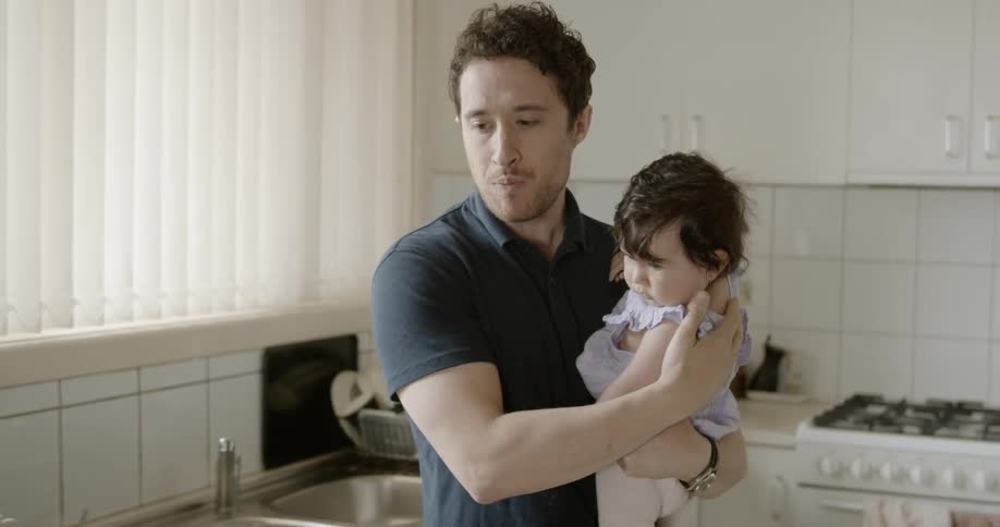 A man standing in a kitchen eating a slice of pastry while holding a baby girl