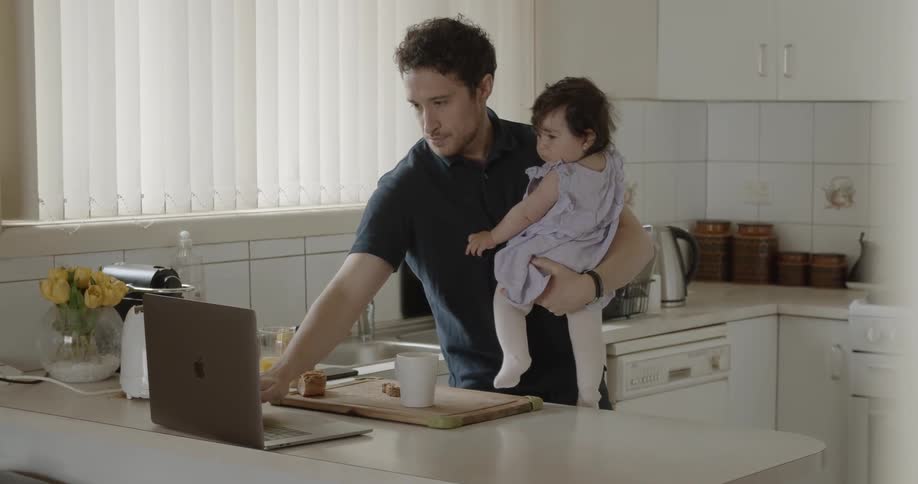 A father holds his infant daughter while standing in the kitchen and typing on his laptop with one hand