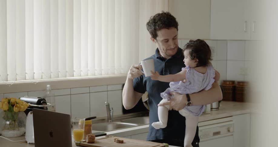 A man takes a sip of coffee while holding a baby girl and watching a laptop in the kitchen
