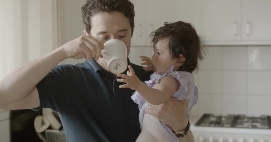 A father drinks coffee while holding his infant daughter in a kitchen