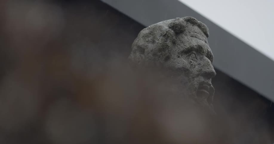 Sleet falls on the bust of a mustachioed man which stands in front of a building