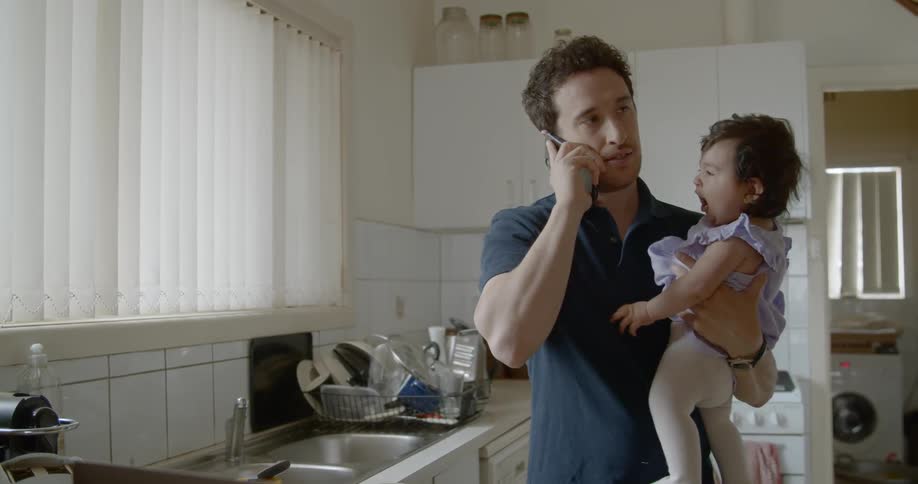 A man answers his phone while holding a baby in the kitchen