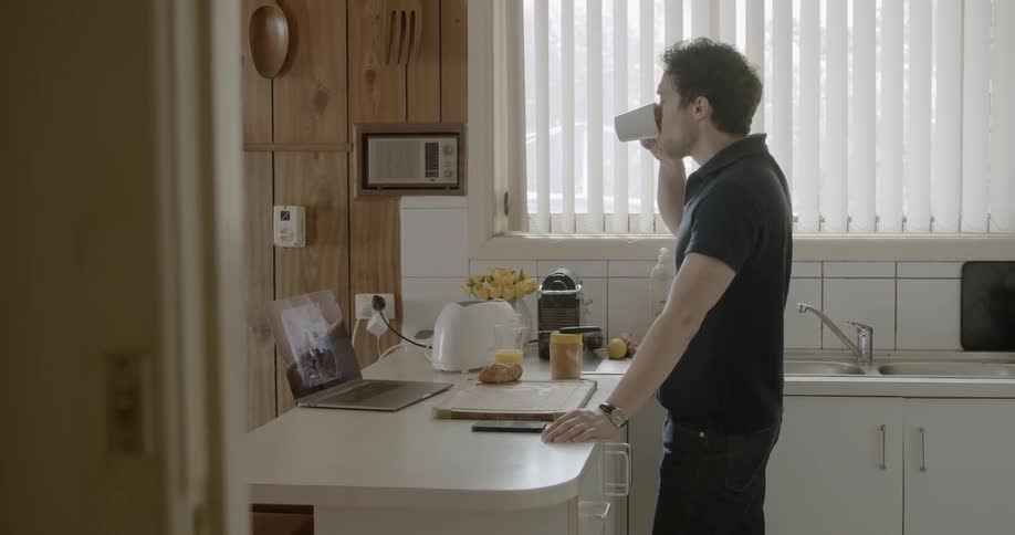 Man watches video from laptop on kitchen the table and sips his coffee in the kitchen during daylight