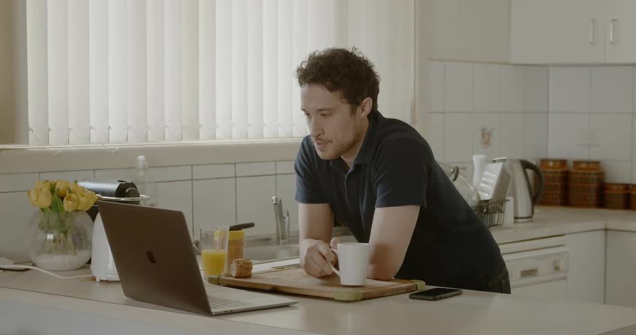 A man is standing hunched over at the kitchen counter drinking from a white mug and working on his laptop