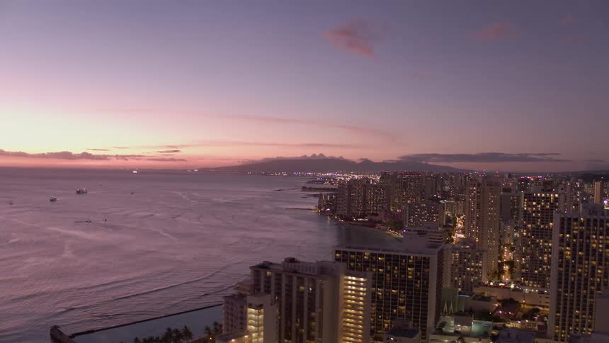 A large downtown area with many tall buildings is illuminated at dusk as this city sits at the edge of the ocean