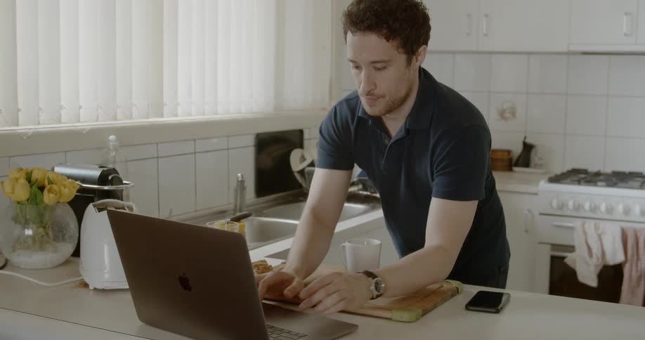 Man puts his elbows on cutting board and uses laptop on the kitchen counter during daylight in the kitchen