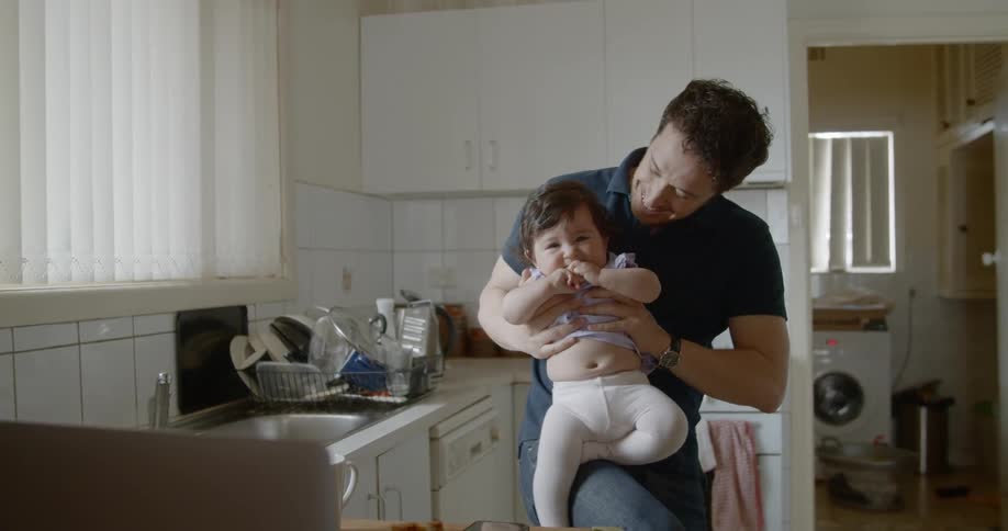 A man in a kitchen holds a baby in his arms trying to get her to smile for a picture