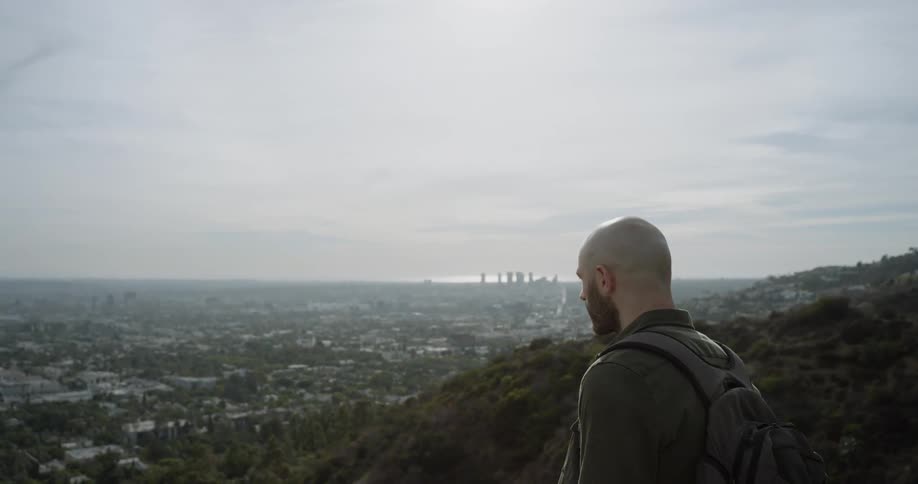 Bald bearded man wearing backpackstands on hill and looks at the distant town during daylight