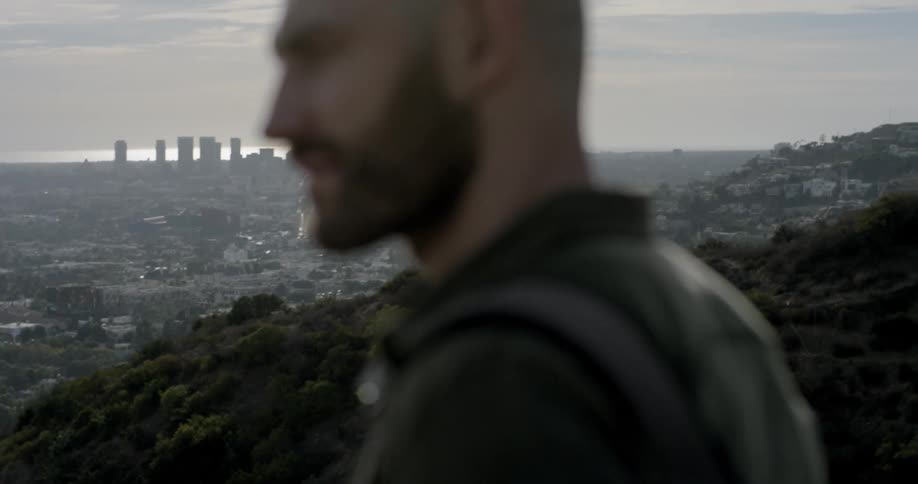 Bald bearded man wearing backpack stands on hill and looks at the city during daylight