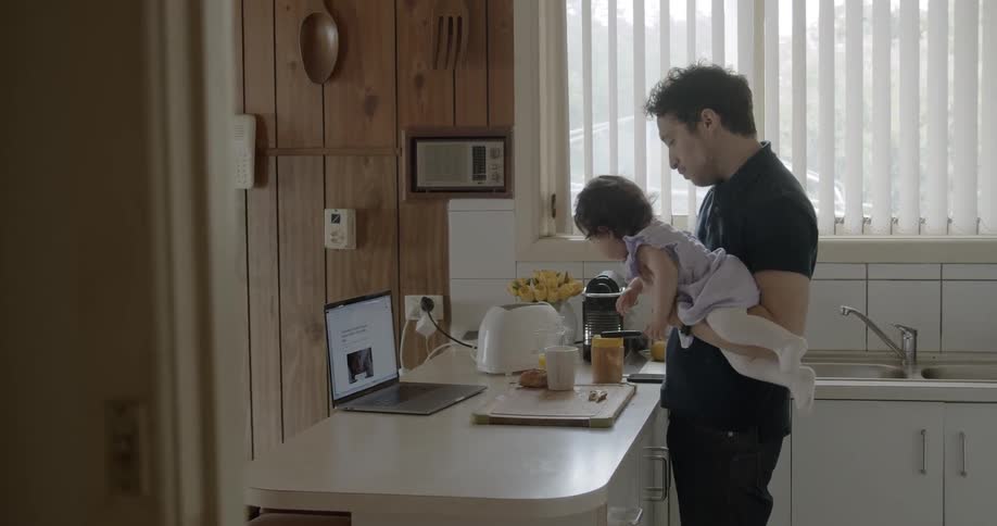 A father holds a baby and feeds her the same thing he is eating while they watch a laptop