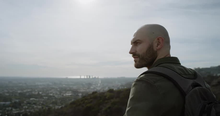 A white man with a beard stands on a hill and looks at a sprawling city