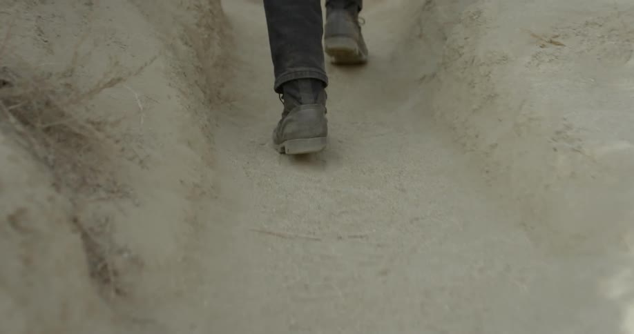 A man wearing black jeans and carrying a backpack climbs a steep dirt trail with unruly shrubs on either side
