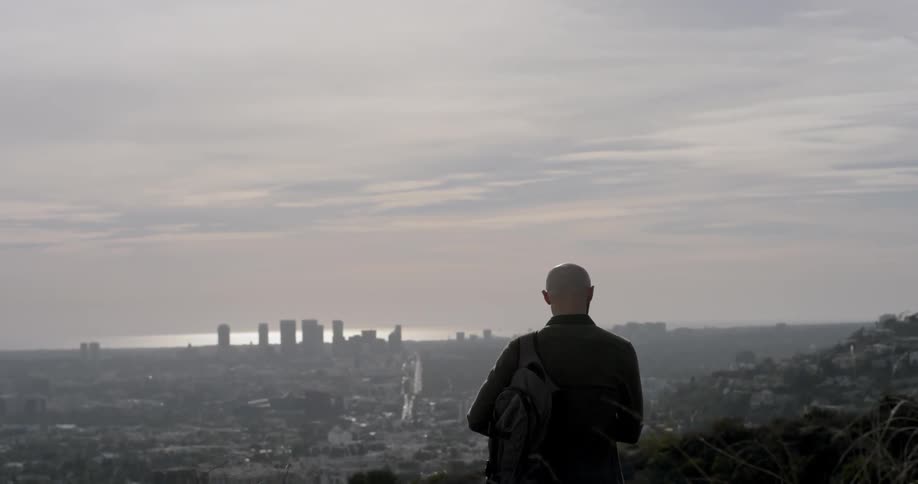 A man with a backpack slung over one shoulder stands on a hill and looks down at the city below and the skyscrapers on the horizon