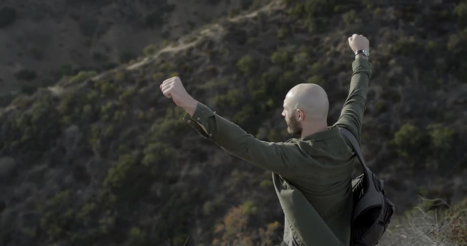 A bald caucasian man raising his arms to the sky while looking down at the landscape in the valley below