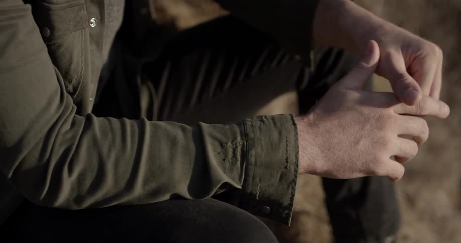 A man sits outside on a hill with his hands clasped together