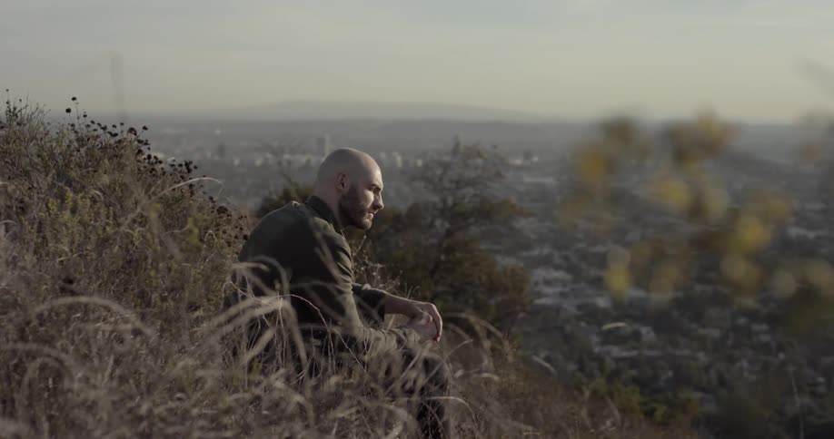 Bald bearded man sits on mountain slope next to the city during daylight