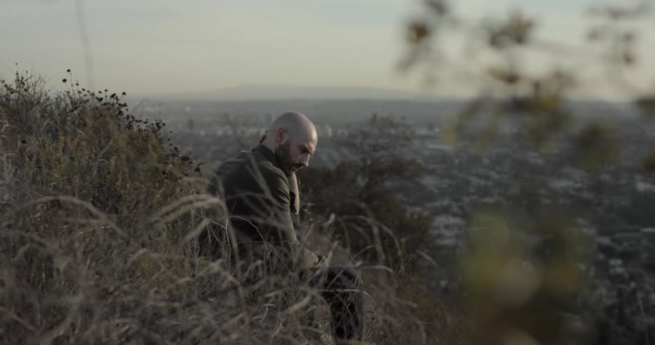 Man with a beard sitting on edge of hillside overlooking the city contemplating life